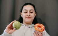 Content young female in hoodie deciding between sweet unhealthy doughnut and green ripe apple in light room