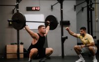 A muscular man performing a weightlifting squat with a coach guiding him indoors.