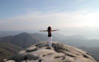 A woman stands and embraces freedom on a mountain peak overlooking a vast landscape.